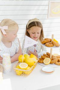 two girls behind a lemonade drink stand with 25 cents sign, a basket of lemons and donuts