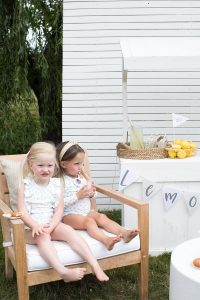 two young girls sitting on a chair in front of a lemonade booth