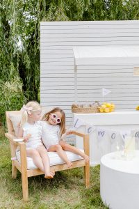 two young girls sitting on a chair in front of a lemonade booth