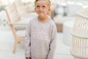 young boy making silly face wearing a gray sun kissed crew sweater standing on a patio