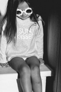 Young Girl wearing sun kissed crew neck sweater and daisy sunglasses sitting on porch