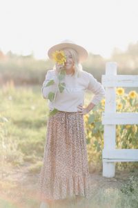 woman wearing a beige hat, country girl sweater and floral print skirt standing in front of a sunflower farm holding a sunflower in front of her face