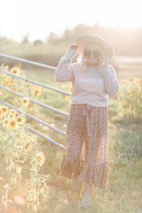 Blonde woman wearing sunglasses hat country girl crew neck sweater floral print skirt walking in a grassy field of sunflowers