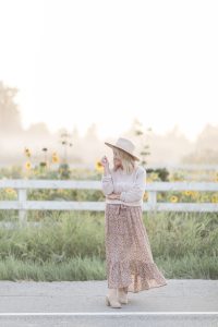 woman wearing a beige hat, country girl sweater and floral print skirt standing in front of a sunflower farm
