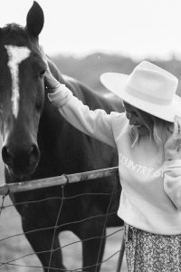 black and white photo woman wearing a hat country girl crew neck sweater and floral print skirt while petting a horse