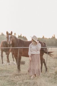woman wearing a beige hat, country girl sweater and floral print skirt standing in front of a horses
