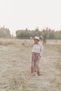 woman wearing a beige hat, country girl sweater and floral print skirt standing in a grassy field