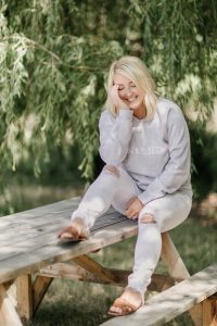 blonde woman smiling sitting on picnic table under a willow tree wearing white ripped jeans with sun kissed sweater
