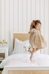 Little Girl Jumping on Bed with #MHxUrbanWalls Brighton Stripes Wallpaper in Vanilla Blush in Background