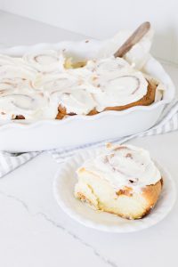 Piece of cinnamon bun with icing on top and a tray of cinnamon buns
