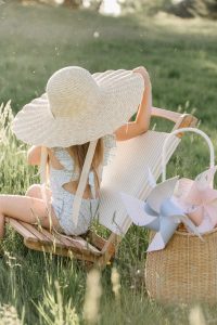 little Girl wearing straw hat and swimsuit sitting on lawn chair next to a basket of pinwheels in grass
